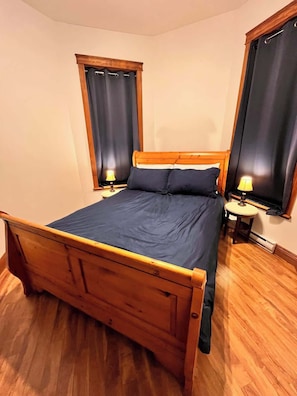 A wooden bed with a blue mattress in a room with wooden flooring, two bedside tables with lamps, and a window with curtains. - Le Juliette  Exceptional site  BBQ & Firepit (Lévis)