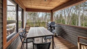 A covered deck with a wooden table and chairs, a grill, and a view of a forest.