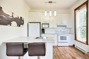 A modern kitchen with white cabinets, a central island with two bar stools, a microwave, a stove, and pendant lighting.