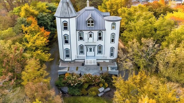 Uma casa grande e branca de dois andares, com um alpendre imponente e telhado inclinado, rodeada por folhagem de outono.