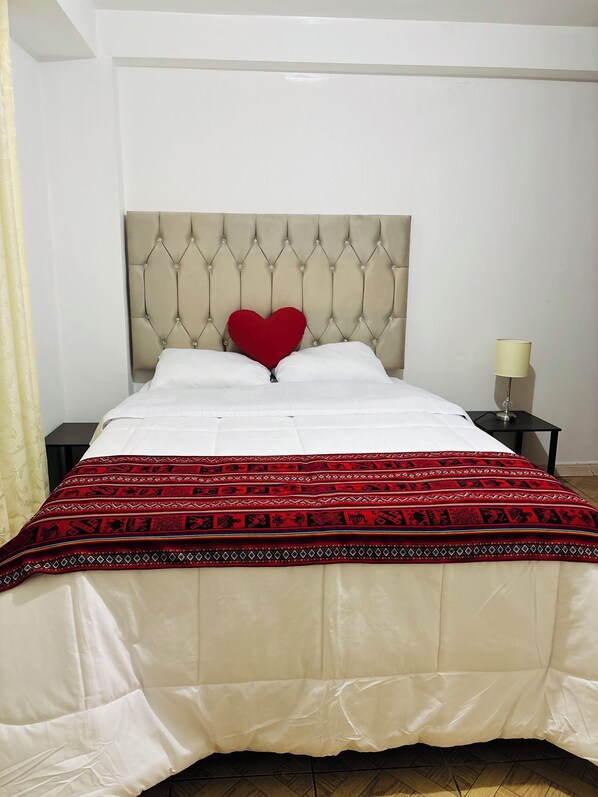 A bed with a tufted headboard, a red heart-shaped pillow, white bedding, and a patterned red and black throw blanket.