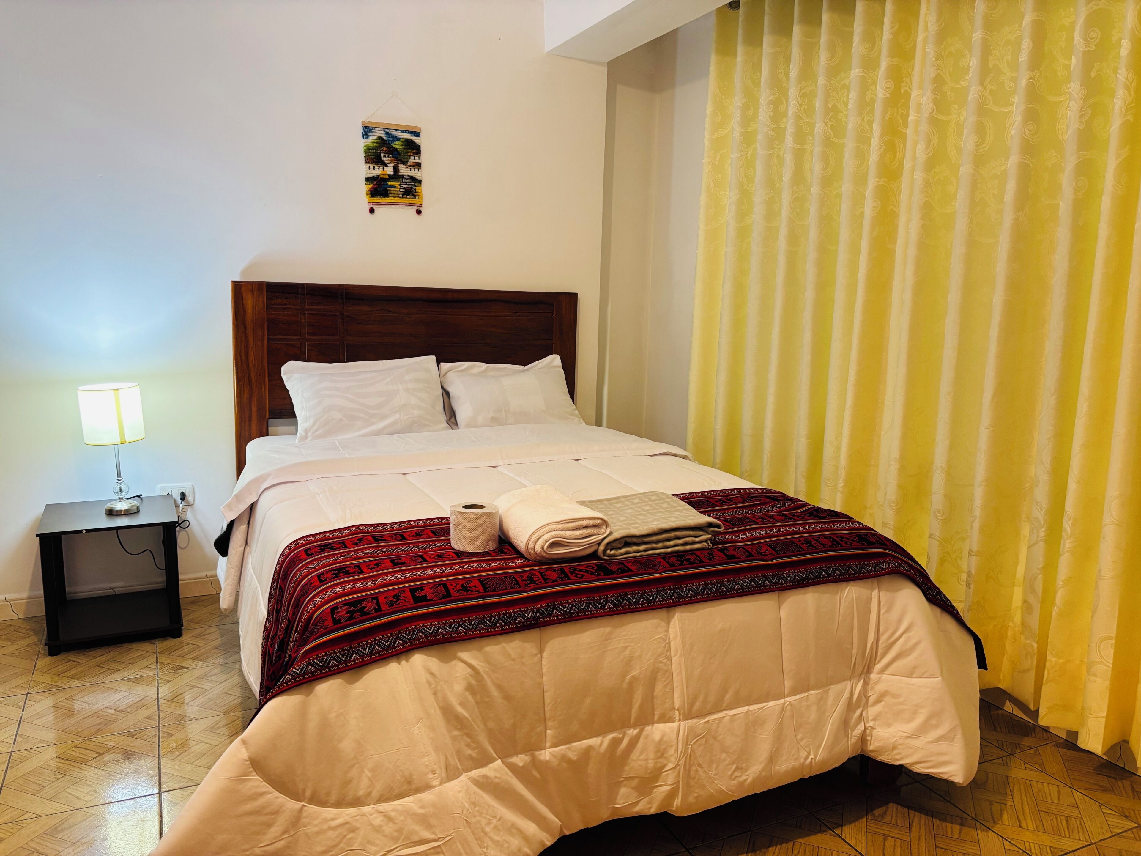 Una cama bien tendida con una colcha de diseño rojo y negro, una mesita de noche con una lámpara y un cuadro colgado en la pared.