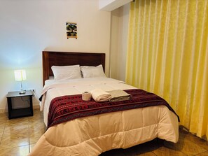 Ein ordentlich bezogenes Bett mit einer rot-schwarz gemusterten Decke, ein kleiner Nachttisch mit Lampe und ein an der Wand hängendes Bild.