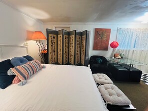 Et hotelværelse med en seng, et glasbord, en stol, en sofa, en lampe, en dekorativ rumdeler og et maleri.