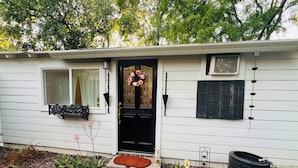 Una casa blanca de un piso con puerta frontal negra, una ventana con cortina blanca, un porche pequeño con cojín rojo y un jardín con plantas y un macetero hecho con un neumático.