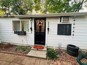 A white single-story house with a black front door, a window with a white curtain, a small porch with a red cushion, and a garden with plants and a tire planter. - Cozy private studio.No pets. Near downtown (Chico)