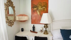 Habitación de hotel con cama, escritorio con silla, mesita de noche, lámpara, espejo, un cuadro y una pequeña estatua.