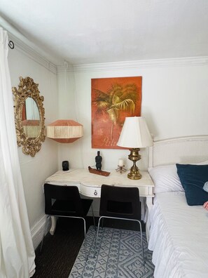 A hotel room with a bed, a desk with a chair, a nightstand, a lamp, a mirror, a painting, and a small statue.
