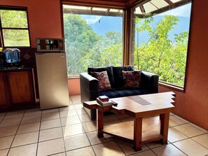 Quarto com sofá, mesa de centro, geladeira e janela com vista para árvores.