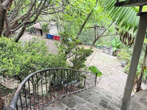 Uma escadaria de pedra leva a uma área coberta com corrimão de metal, cercada por vegetação e um pequeno edifício com lixeiras coloridas.