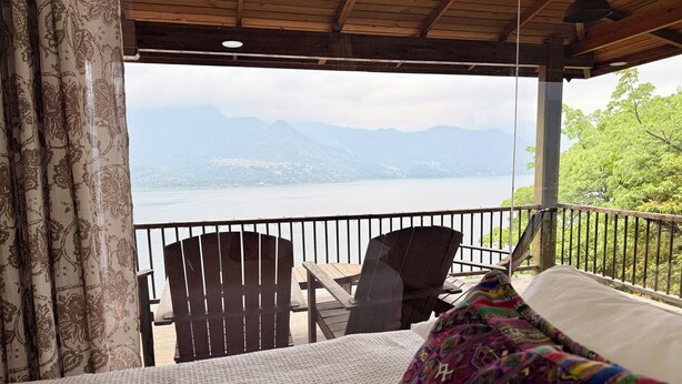 A room with a bed, a view of a lake and mountains, a wooden ceiling, and a balcony with chairs. - Apartment # 1 Chi-ya, San Juan laguna (San Juan la laguna)