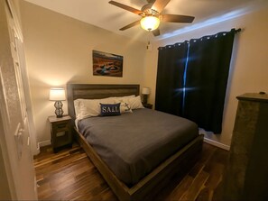 Une chambre à coucher avec un lit, un ventilateur de plafond, un tableau, une table de chevet, une lampe et une fenêtre avec des rideaux.