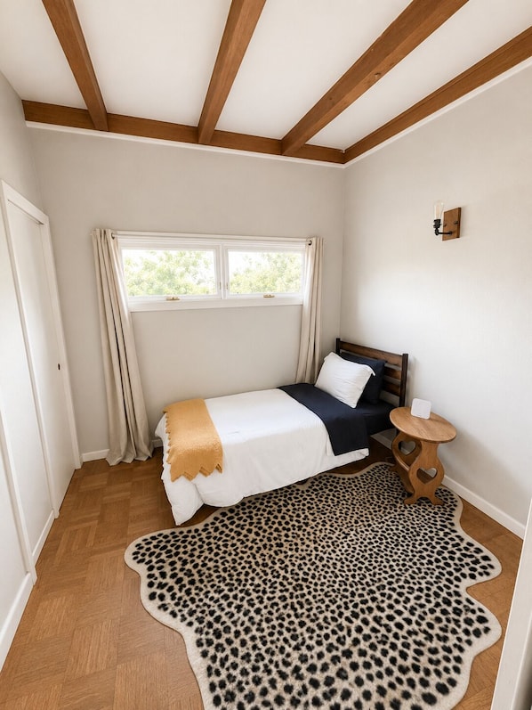 A bedroom with a bed, a window, a wooden ceiling, a small round table, and a leopard print rug.