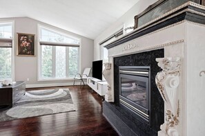 Una habitación con chimenea, un ventanal grande, una silla blanca y un televisor.