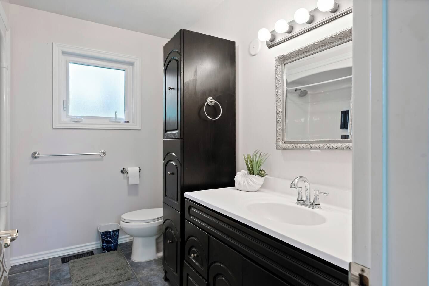 A bathroom with a white vanity, a dark cabinet, a mirror with lights, a window, and a toilet.