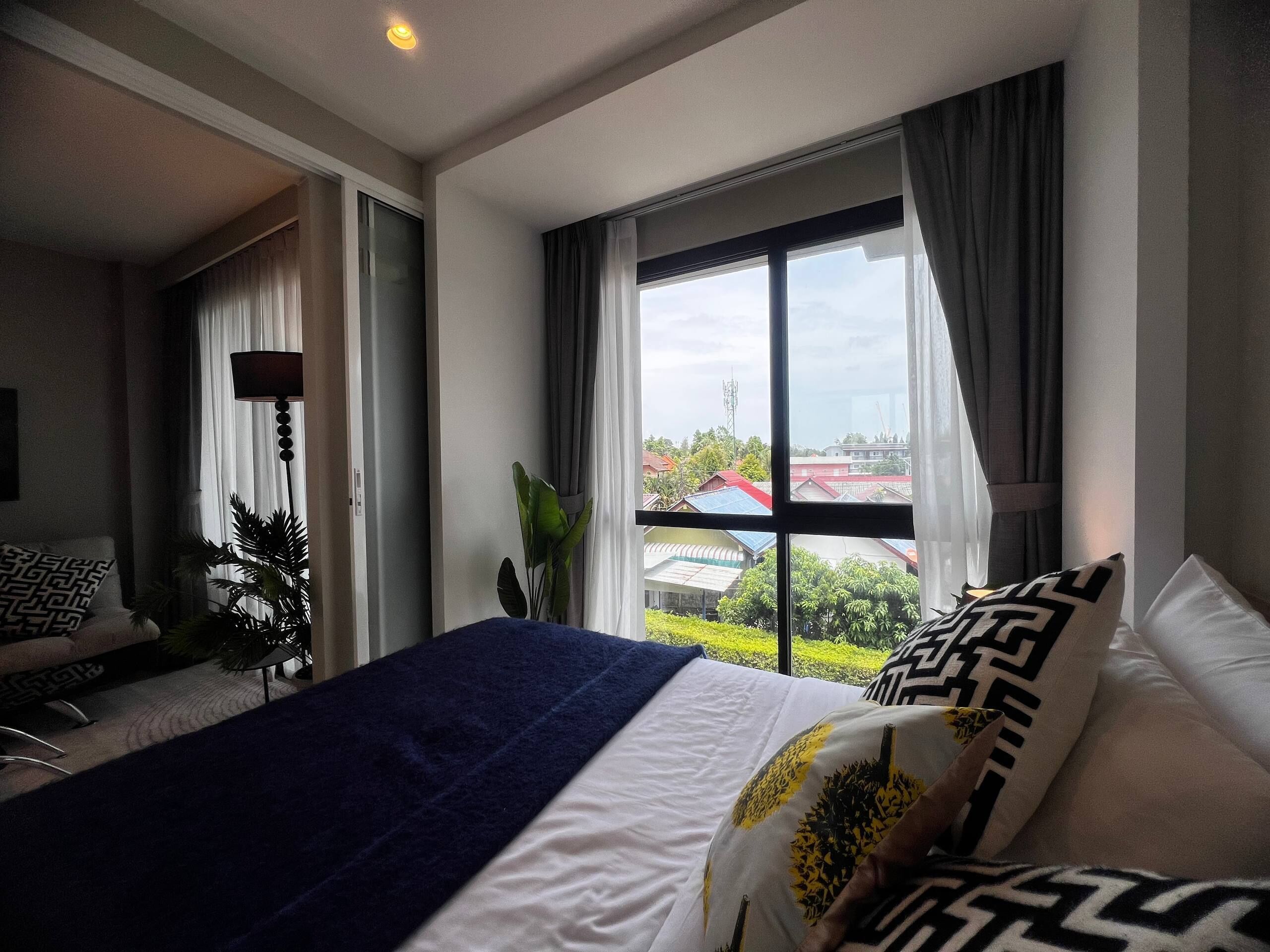 A hotel room with a bed, a desk, a lamp, and a view of buildings through large windows.