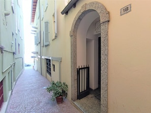 A narrow alleyway with a potted plant, a number 18 sign, and a door with a black gate. - Antica_Stresa_1_apartment (Stresa)