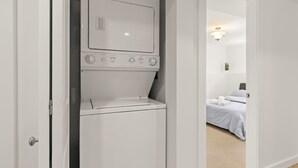Uma lavanderia compacta e branca, com máquina de lavar e secadora embutidas, uma cama com roupa de cama branca e uma pequena planta na mesinha de cabeceira.
