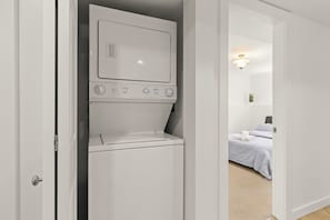 Ein kleiner, weißer Hauswirtschaftsraum mit integrierter Waschmaschine und Trockner, einem Bett mit weißer Bettwäsche und einer kleinen Pflanze auf dem Nachttisch.