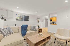 Ein Wohnzimmer mit einer Couch, einem Couchtisch und einem Sessel. Durch eine offene Tür ist eine Küche zu sehen.