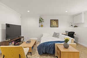 Ein modernes Wohnzimmer mit einem Flachbildfernseher, einem Holztisch, einer Couch, einem Bett mit blauer Decke und einem Schreibtisch mit einem Laptop.