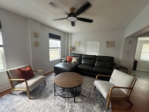 Ein Wohnzimmer mit einem schwarzen Ledersofa, zwei Sesseln, einem runden Couchtisch und einem Deckenventilator.