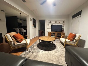 Una sala de estar con dos sillones, una mesa redonda de centro, un televisor sobre un mueble bajo, y un comedor al fondo.