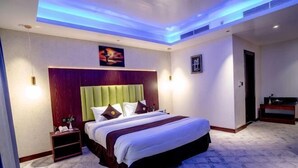 A hotel room with a bed, bedside table, nightstand, and a painting on the wall. - Raha Grand Hotel (Dubai)