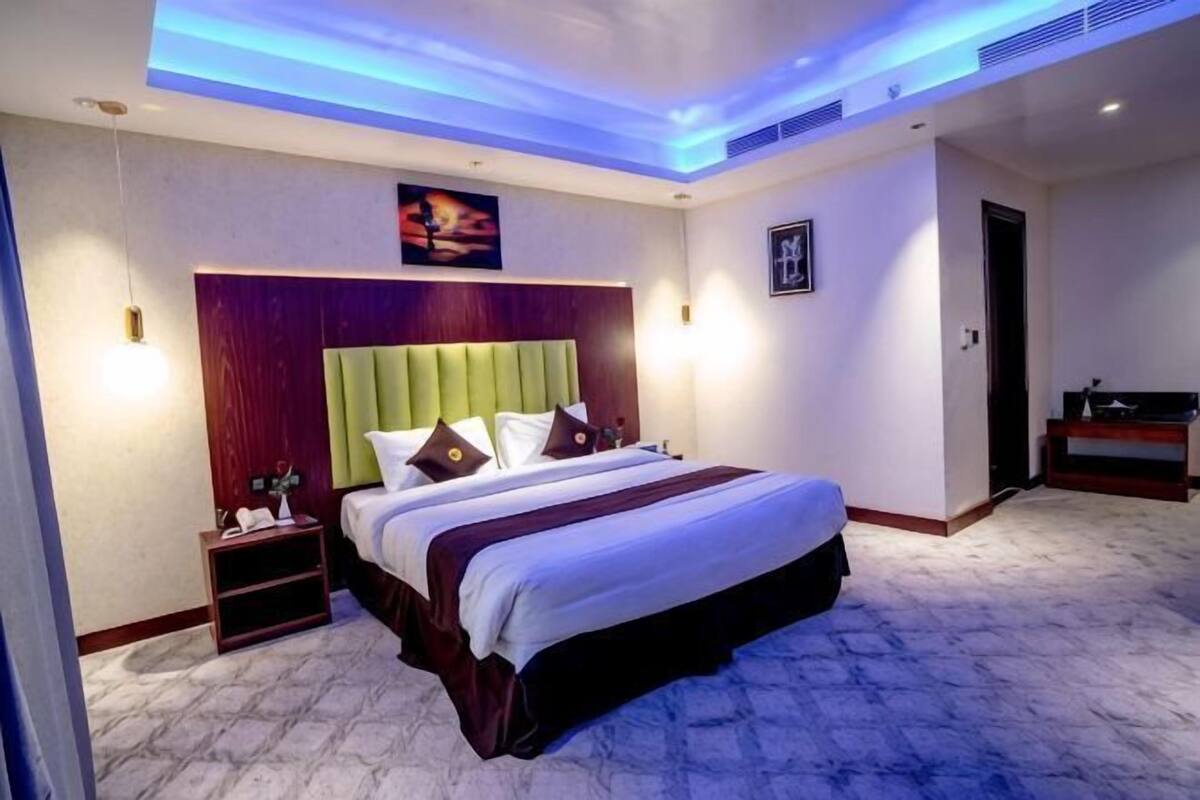 A hotel room with a bed, bedside table, nightstand, and a painting on the wall.