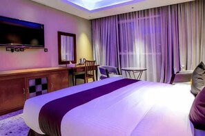 A hotel room with a large bed, a desk with a chair, a television, and a window with curtains. - Raha Grand Hotel (Dubai)