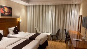A hotel room with two beds, a desk, a chair, a TV, and a large window with curtains. - Raha Grand Hotel (Dubai)