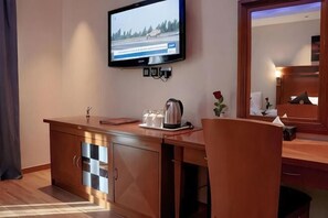 A hotel room with a flat-screen TV mounted on the wall, a wooden desk with a coffee maker, a mirror, and a small plant. - Raha Grand Hotel (Dubai)