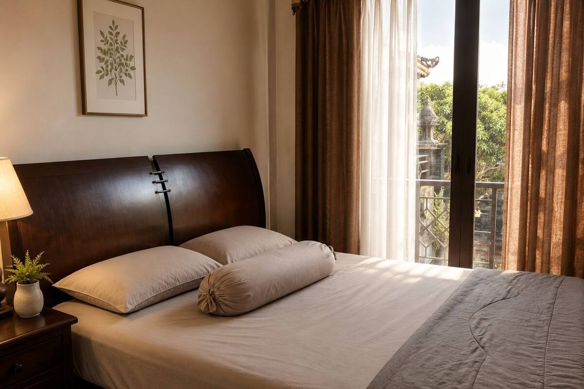 A neatly made bed with a headboard, a bedside table with a lamp and a plant, and a window with curtains.