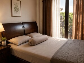 Una cama bien hecha con cabecera, una mesita de noche con lámpara y planta, y una ventana con cortinas.