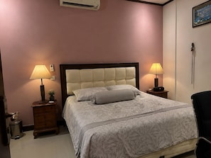 A bedroom with a bed, two bedside tables with lamps, a wall-mounted air conditioner, and a painting on the wall. - Griyaku Homestay (Badung)