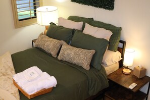 Een net opgemaakt bed met groene beddengoed, een bedlampje en een mandje met opgevouwen handdoeken.