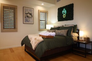 Una camera da letto con un letto, un comodino, una lampada, una decorazione a forma di cactus appesa al muro e due finestre con persiane.
