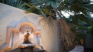 Ein Steinkonstrukt mit einer Fontäne, aus deren Zentrum Wasser sprüht, umgeben von einer niedrigen Ziegelmauer und Palmen.