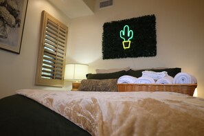 A bedroom with a bed, a lamp, a wall-mounted cactus decoration, and a basket of towels. - Stylish Studio on Roosevelt Row – Fast Wi-Fi (Phoenix)