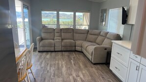 A room with a sectional sofa, a dining table with chairs, a kitchen area, and large windows with a view of mountains. - Relaxing 2 Bedroom with Panoramic Lake View from inside and out (Lake Havasu City)