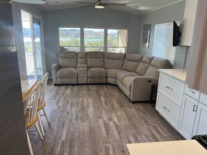 Ein Zimmer mit Ecksofa, einem Esstisch mit Stühlen, einer Küchenzeile und großen Fenstern mit Bergblick.