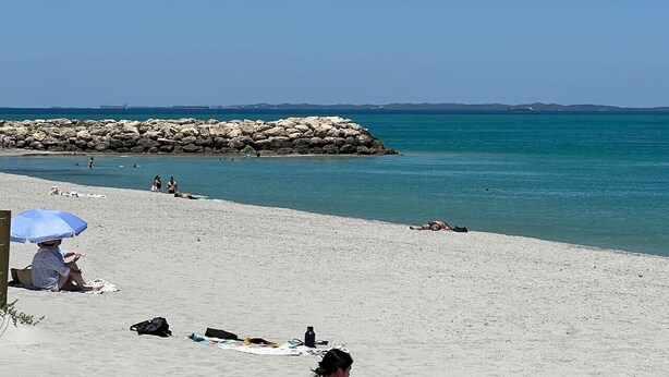 Uma praia com pessoas tomando sol, um molhe rochoso e mar calmo.