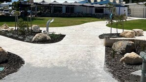 Um edifício moderno com área externa coberta, caminho de concreto e canteiros de jardim ajardinados com pedras.
