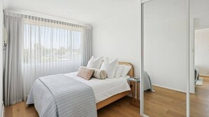 Quarto com cama, mesa de cabeceira, espelho e janela com cortinas leves.