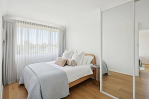 Een slaapkamer met een bed, nachtkastje, spiegel en raam met dunne gordijnen.