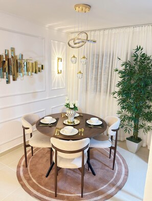 Ein Esszimmer mit einem runden Tisch, Stühlen, einer Vase mit Blumen, einer Topfpflanze und einer Wandleuchte.