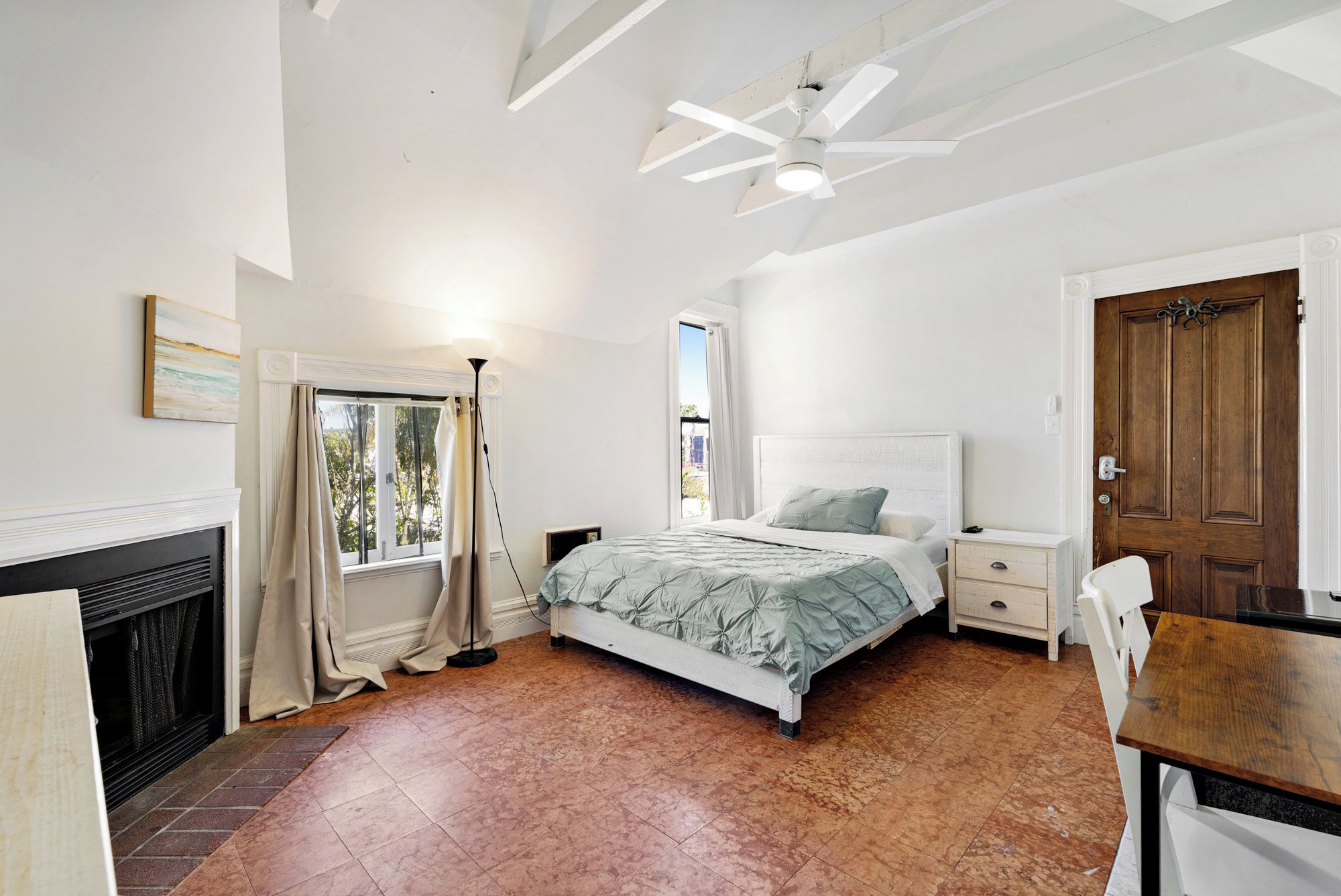 A bedroom with a bed, bedside tables, a wooden door, a window with curtains, a ceiling fan, and a fireplace.