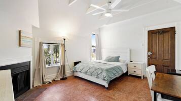 A bedroom with a bed, bedside tables, a wooden door, a window with curtains, a ceiling fan, and a fireplace.