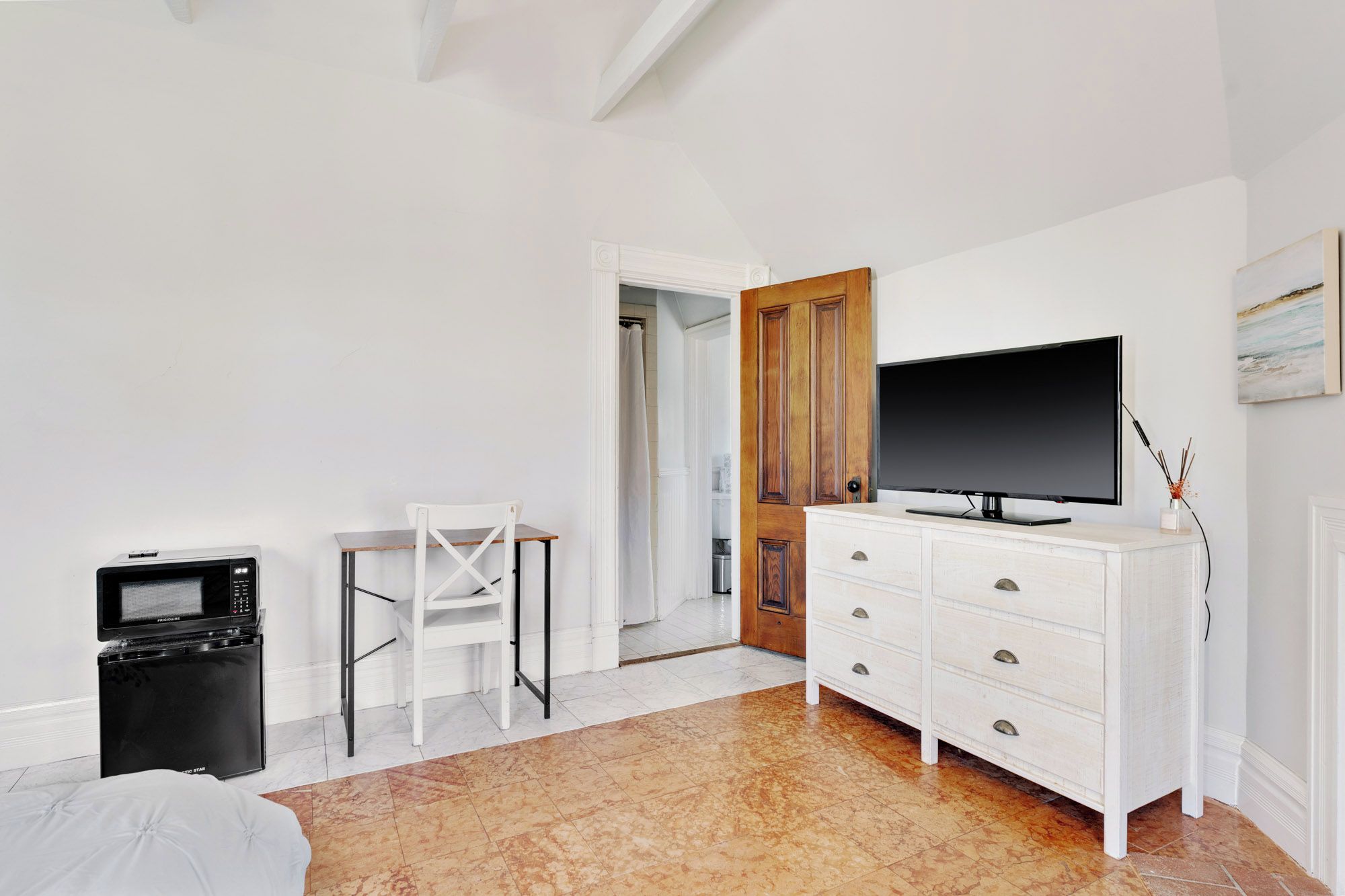 A bedroom with a television on a dresser, a microwave, a chair, and a door leading to another room.