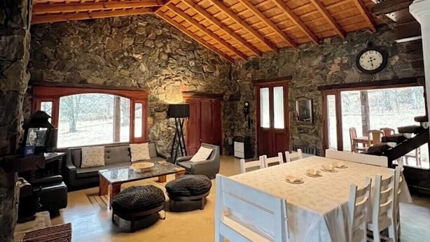 Ein rustikales Interieur mit einer Steinwand, Holzbalkendecke, einem für sechs Personen gedeckten Esstisch und einem Kamin.